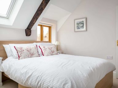 A bedroom with a double bed white bedding and decorative pillows wooden window and exposed beam at Oyster Barn in Naunton