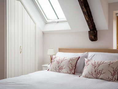 A bedroom with a bed with patterned pillows a bedside table with a lamp a skylight window and a wooden beam at Oyster Barn in Naunton