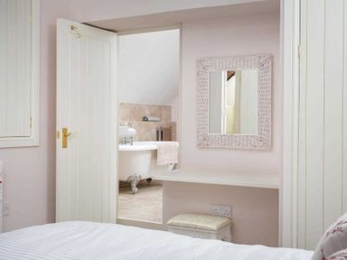 A bedroom with a door open to a bathroom showing a clawfoot bathtub a mirror above a desk and a stool under the desk at Oyster Barn in Naunton