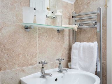 A bathroom sink with two taps a glass shelf with cups and soap and a towel on a radiator at Oyster Barn in Naunton