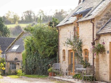 An outdoor seating area with metal chairs and a table beside a stone building with plants and trees around at Oyster Barn in Naunton