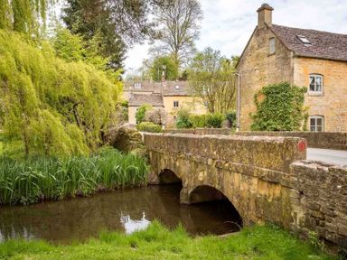 A stone bridge over a small waterway with greenery and old stone buildings at Oyster Barn in Naunton