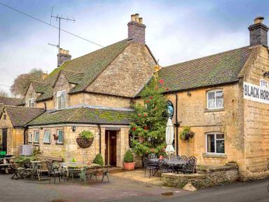 An outdoor seating area with tables and chairs in front of a stone building with a sign that says The Black Horse at Oyster Barn in Naunton