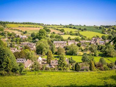 A village with houses and trees surrounded by green fields under a blue sky at Oyster Barn in Naunton