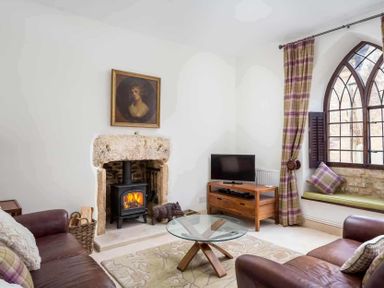 A living room with a fireplace a wood stove two leather sofas a glass coffee table a television on a wooden stand and a window seat with cushions at Little Culver in Minchinhampton