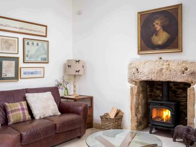 A living room with a brown leather sofa pillows a wood burning stove in a stone fireplace and framed pictures on the wall at Little Culver in Minchinhampton