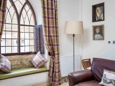 A seating area with a window seat with cushions and plaid curtains next to a floor lamp and a leather sofa at Little Culver in Minchinhampton
