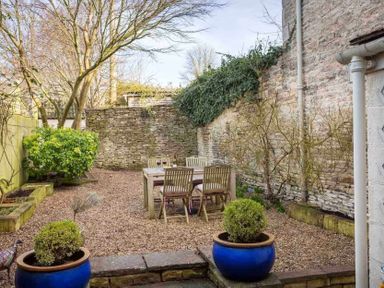 An outdoor garden area with a wooden table and chairs on gravel surrounded by stone walls and plants at Little Culver in Minchinhampton