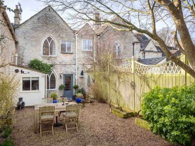 An outdoor patio area with a wooden table and chairs gravel ground and a stone building in the background at Little Culver in Minchinhampton