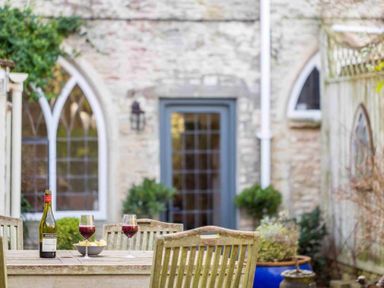 An outdoor wooden table with two glasses of red wine a bowl of chips and a bottle of wine in a garden at Little Culver in Minchinhampton