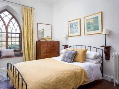 A bedroom with a bed with yellow bedding and pillows a wooden dresser a mirror two framed pictures and a window with yellow curtains at Little Culver in Minchinhampton