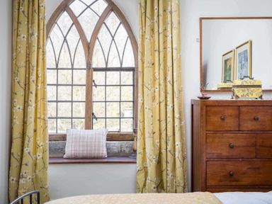 A bedroom with a wooden dresser a mirror floral curtains and a pillow on a window ledge at Little Culver in Minchinhampton