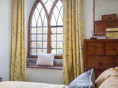 A bedroom window with floral yellow curtains and a plaid pillow on the windowsill next to a wooden dresser with a mirror at Little Culver in Minchinhampton