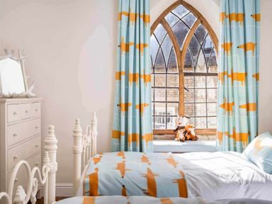 A bedroom with twin beds blue and orange fox patterned bedding and curtains a wooden arched window a white dresser and stuffed animals on the window seat at Little Culver in Minchinhampton