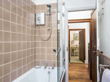 A bathroom with a bathtub shower and beige tiled walls leading to a hallway and a room with a desk and chair at Little Culver in Minchinhampton