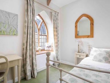 A bedroom with a bed, bedside table and lamp, a desk with a chair, arched window with curtains, and a wooden framed mirror at Little Culver in Minchinhampton