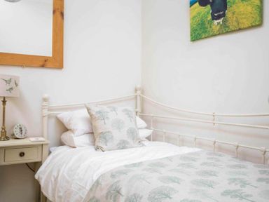 A bedroom with a white metal framed bed a bedside table with a lamp and clock and a painting on the wall at Little Culver in Minchinhampton