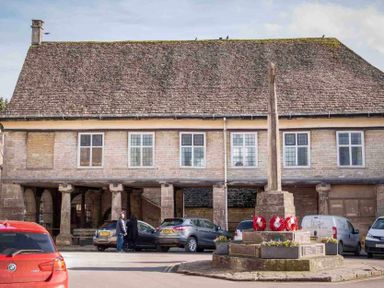 A street scene with parked cars two people and a stone building with columns and a monument in front at Little Culver in Minchinhampton