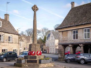 A stone war memorial with wreaths in a village square surrounded by stone buildings and parked cars at Little Culver in Minchinhampton