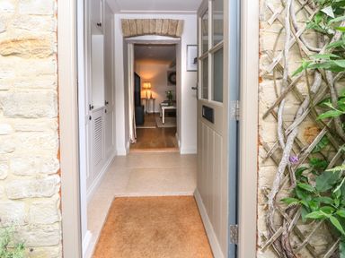 Open front door leading to hallway with built-in white cabinets and view into living area with lamp and table at Spinners Cottage in Stow-on-the-Wold