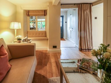 A sitting area with a beige sofa and side table with a lamp and decorative items and a window seat next to a passage with a curtain and a glass coffee table with magazines and flowers at Spinners Cottage in Stow-on-the-Wold
