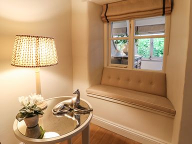 A corner with a small round glass table holding a lamp flower pot and dog figurine next to a cushioned window seat at Spinners Cottage in Stow-on-the-Wold
