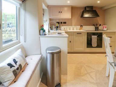 A kitchen with beige cabinets stainless steel oven and a built in hob a window seat with a cushion and a stainless steel bin at Spinners Cottage in Stow on the Wold