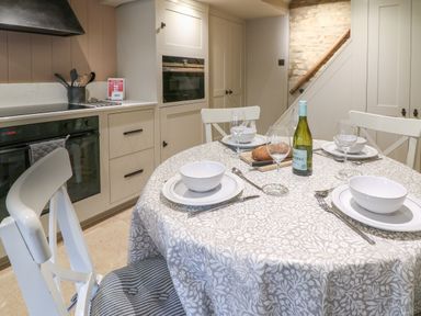 A kitchen with a round dining table set for four with plates bowls wine glasses and a bottle of wine at Spinners Cottage in Stow-on-the-Wold