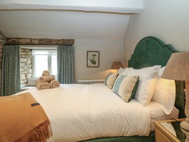 A bedroom with a green headboard bed white bedding rolled towels on bed green patterned cushions bedside table with lamp and stone windowsill at Spinners Cottage in Stow-on-the-Wold