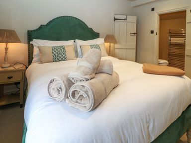 A bedroom with a green upholstered headboard a bed with white bedding and rolled towels two bedside tables with lamps and an open door leading to a bathroom at Spinners Cottage in Stow-on-the-Wold