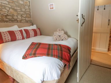 A bedroom with a bed made with white bedding red pillows and a folded red plaid blanket with rolled towels on top at Spinners Cottage in Stow-on-the-Wold