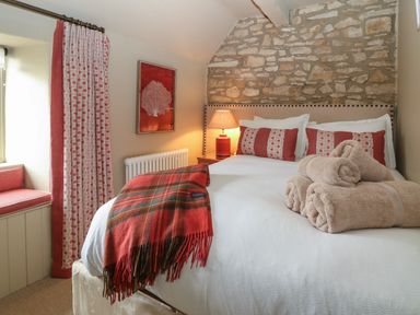 A bedroom with a stone wall headboard a bed with red cushions a red plaid blanket beige towels a bedside lamp and red patterned curtains at Spinners Cottage in Stow-on-the-Wold