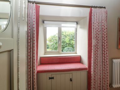 A window seat with red cushion and patterned curtains by a window showing greenery outside at Spinners Cottage in Stow-on-the-Wold