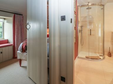 A partially open door showing a bedroom with a window seat and curtains on the left and a bathroom with a glass shower enclosure on the right at Spinners Cottage in Stow-on-the-Wold