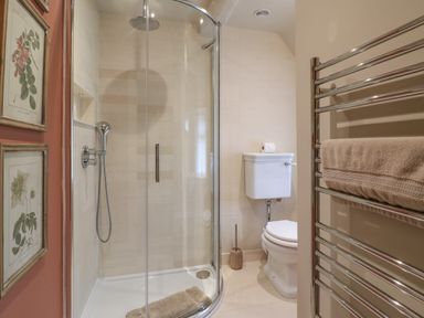 A bathroom with a curved glass shower enclosure a toilet a towel rack with a beige towel and botanical prints on the wall at Spinners Cottage in Stow-on-the-Wold
