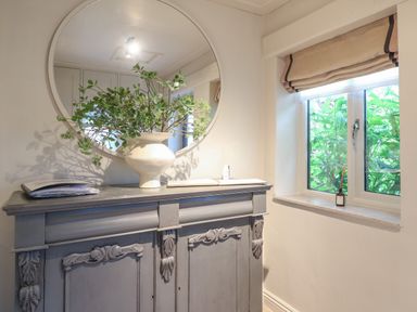 A wooden cabinet with decorative carvings a large round mirror a vase with green foliage on the cabinet and a window with beige roman blinds at Spinners Cottage in Stow-on-the-Wold