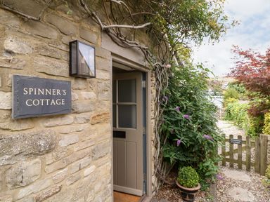 The entrance of a stone cottage with an open door a wall sign reading Spinners Cottage plants and a small wooden gate at Spinners Cottage in Stow-on-the-Wold
