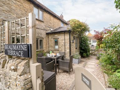 Exterior of a stone cottage with a garden path and outdoor dining table with chairs at Spinners Cottage in Stow-on-the-Wold