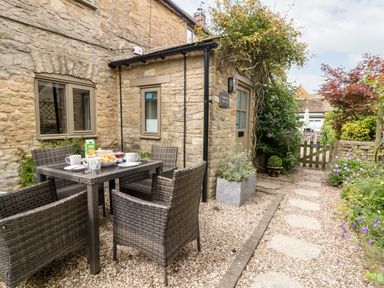 A stone cottage with an outdoor dining table and chairs on a gravel patio surrounded by plants at Spinners Cottage in Stow on the Wold