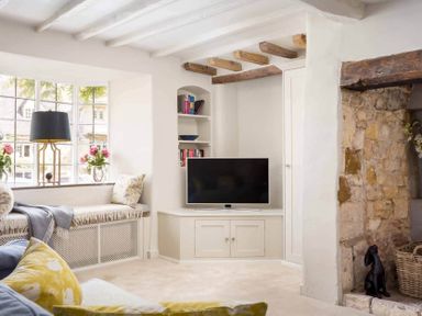 A living room with a window seat flowers on the window ledge a television built into a white cabinet exposed wooden ceiling beams and a stone fireplace at Stanley Cottage in Chipping Campden