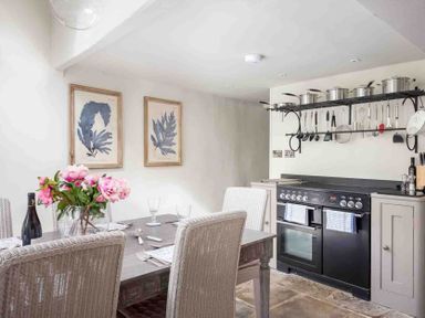 A kitchen with a black stove and hanging pots above a wooden dining table with pink flowers at Stanley Cottage in Chipping Campden
