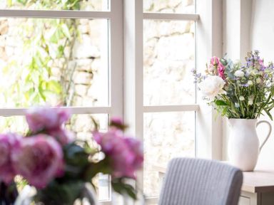 A table with a white pitcher holding flowers and a window with plants outside at Stanley Cottage in Chipping Campden