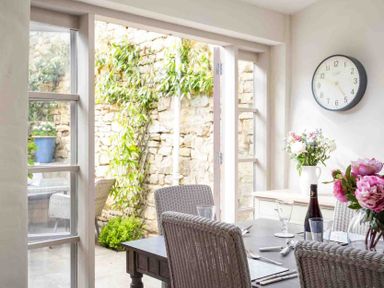 A dining area with wicker chairs a table set with glasses cutlery and flowers and a wall clock at Stanley Cottage in Chipping Campden