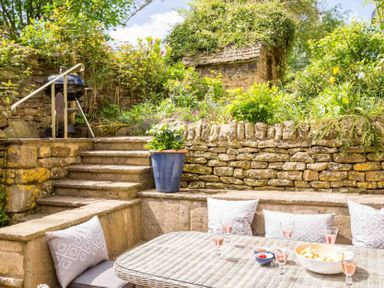A garden seating area with cushions and drinks on a table next to stone steps and a barbecue at Stanley Cottage in Chipping Campden
