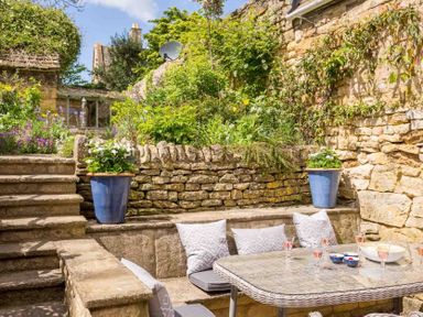 An outdoor stone patio area with built-in seating and a glass table with cushions and plants at Stanley Cottage in Chipping Campden