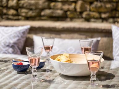 A glass table with four glasses of pink drink a bowl of chips and three small bowls with snacks and pillows against a stone wall at Stanley Cottage in Chipping Campden