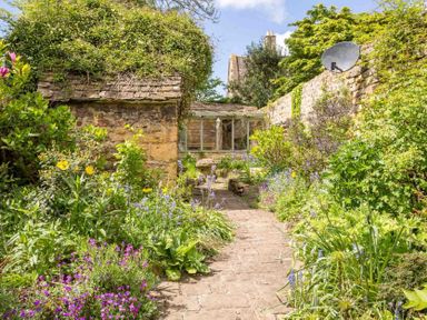 A garden path with flowering plants on both sides and a stone building with a tiled roof at Stanley Cottage in Chipping Campden