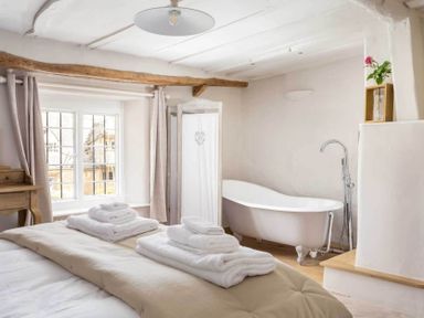 A bedroom with a bed folded towels a clawfoot bathtub and a window with curtains at Stanley Cottage in Chipping Campden