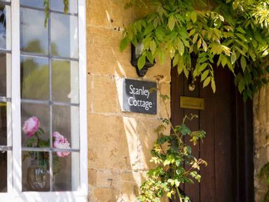 A cottage entrance with a wooden door a sign saying Stanley Cottage and flowers visible through a window at Stanley Cottage in Chipping Campden