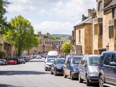 A street with parked cars and stone buildings with a tree on the left side at Stanley Cottage in Chipping Campden